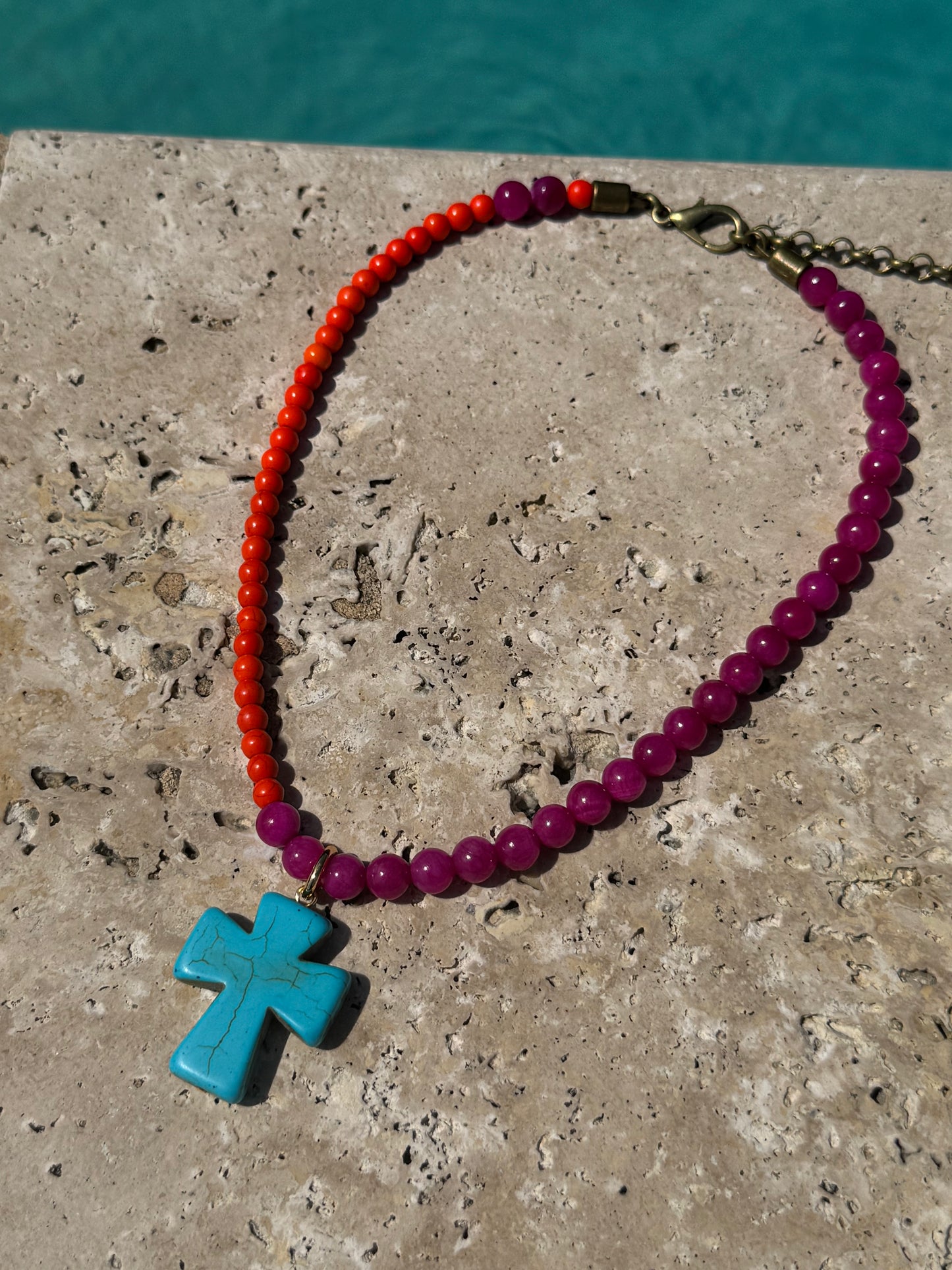 Necklace with red and purple beads and a blue cross pendant on a stone surface with water in the background