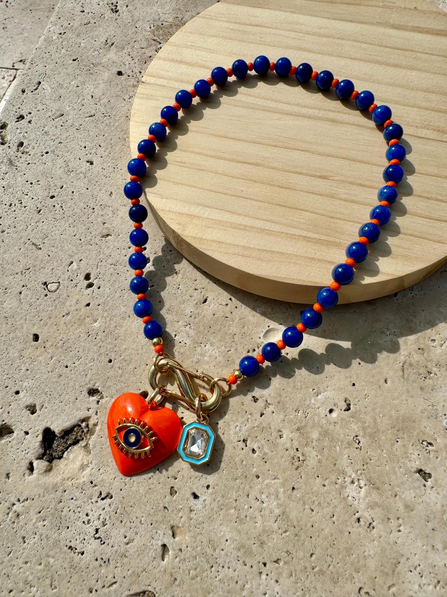May include: A necklace with blue and orange beads and a gold clasp. The necklace has a heart-shaped charm with an evil eye design and a small diamond-shaped charm.