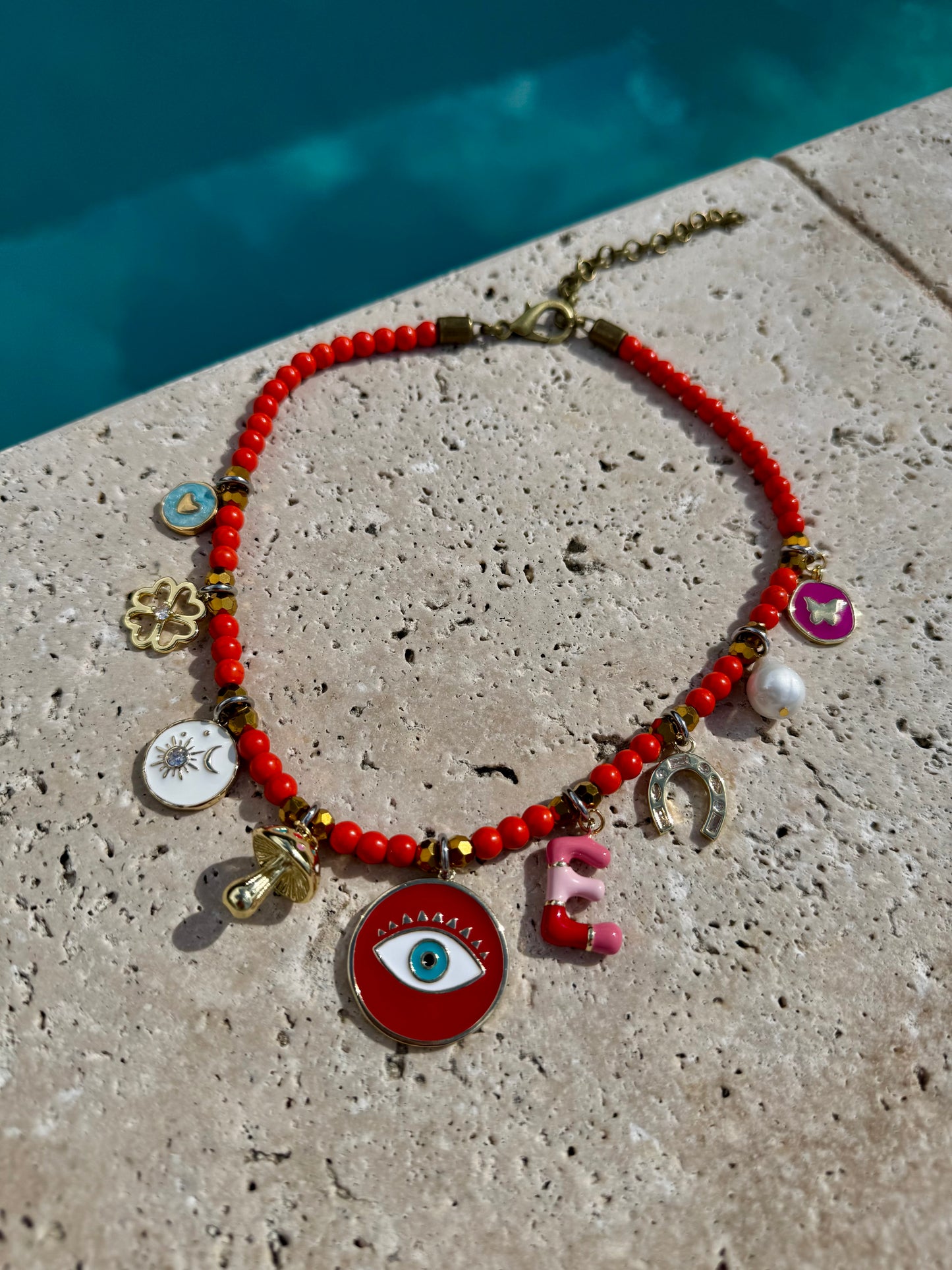Necklace with red beads and various charms on a stone surface near a pool.
