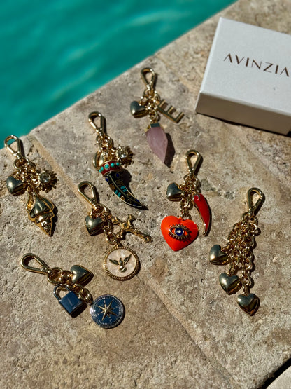 Gold charm keychains with various charms on a stone surface next to an Avinzia box.