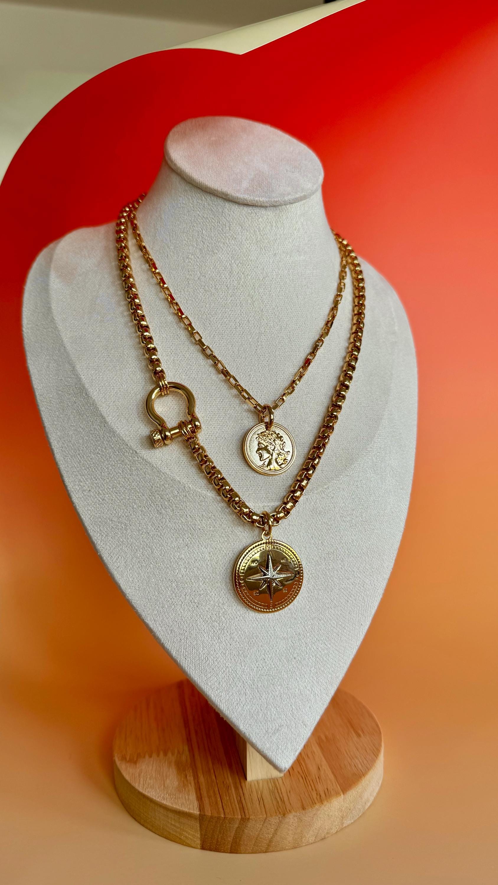 A gold necklace with two pendants, one featuring a compass design and the other a round coin with a profile image. The necklace is displayed on a white velvet heart-shaped stand on a wooden base.