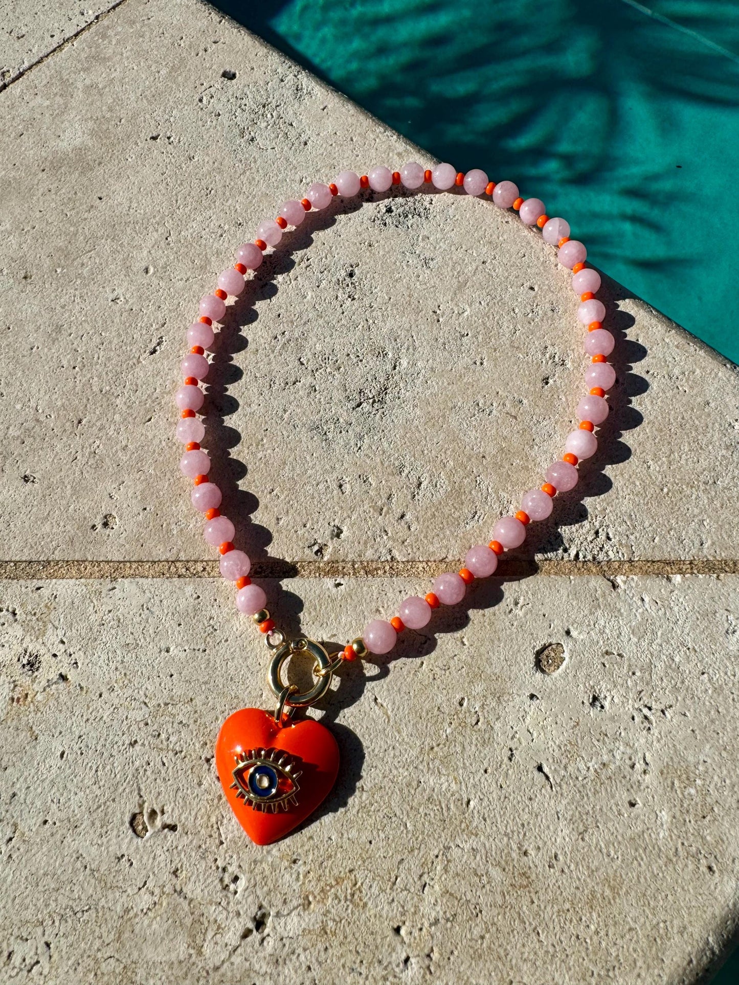 May include: A pink and orange beaded necklace with a red heart charm featuring a gold evil eye.